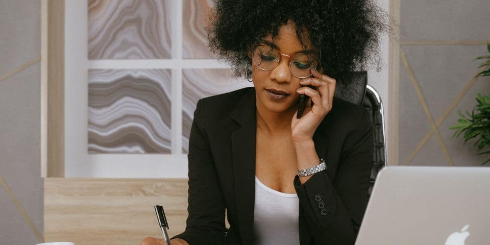 black woman sitting at a desk while on the phone and writing something with a pen