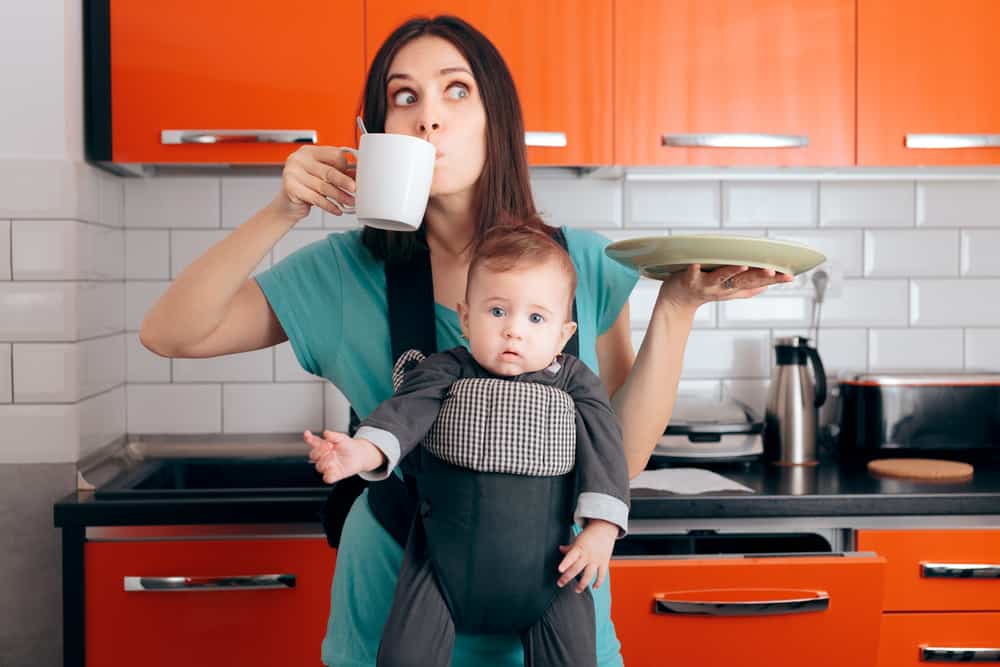 overwhelmed white mom with a baby in a baby carrier strapped to her chest drinking coffee and holding a plate