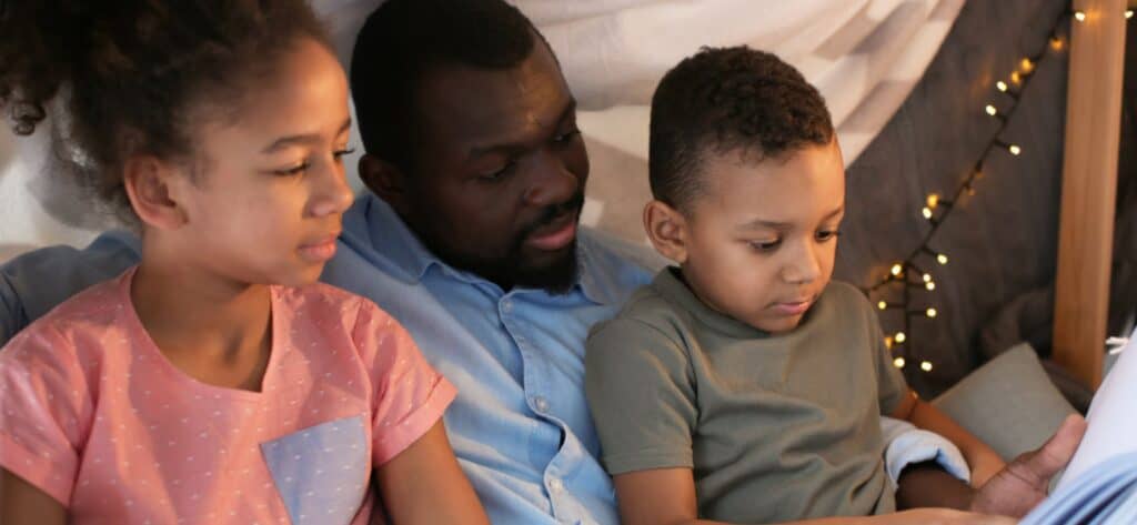 black dad, daughter and son reading a book in a bed tent with lights before bed