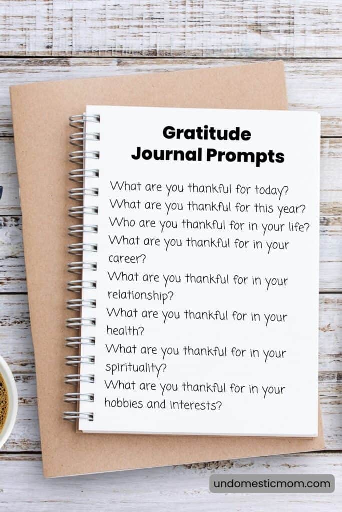 An open notebook stacked on a brown book that says "gratitude journal prompts" and lists ten prompts