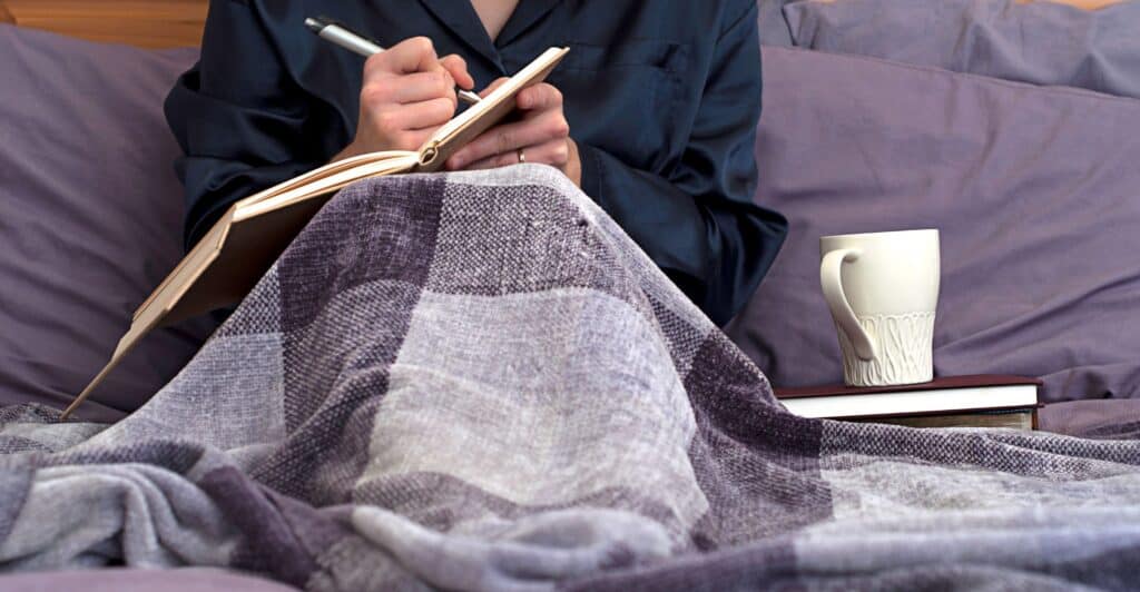 A caucasian person in bed, not showing their head, journaling with a cup of tea next to them.